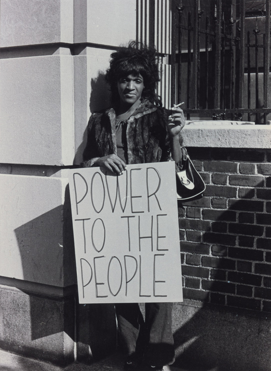 Marsha P. Johnson standing outside of a hospital with a sign that reads 'Power to the People